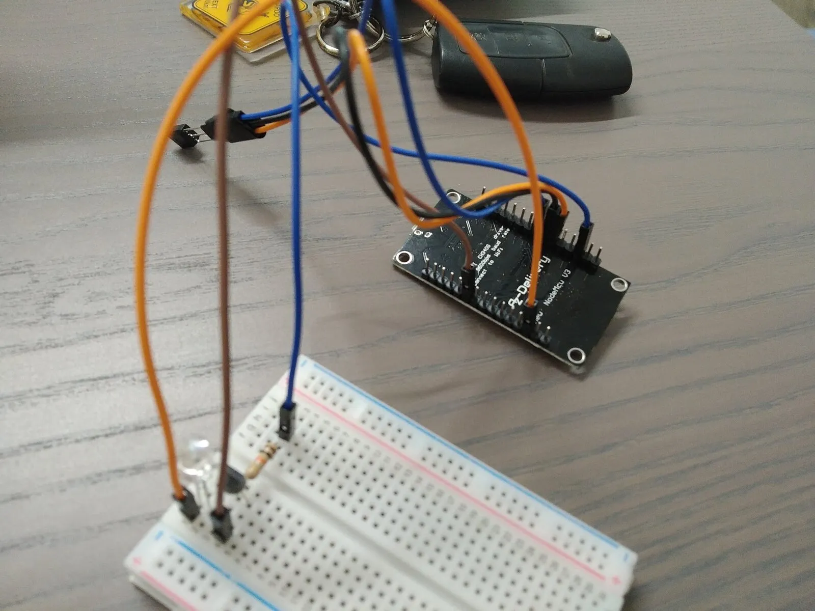 A breadboard on a desk holding a clear infrared LED, a small black transistor and a resistor, wired with orange and blue jumper leads to an ESP8266 development board lying beside it